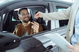 Homem sentado dentro do carro recebe a chave do veículo novo com expressão de felicidade