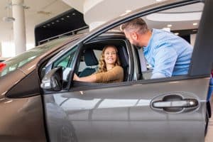 Smiling mid-adult couple buying a new car from dealership, woman sitting inside the vehicle while man standing beside her.